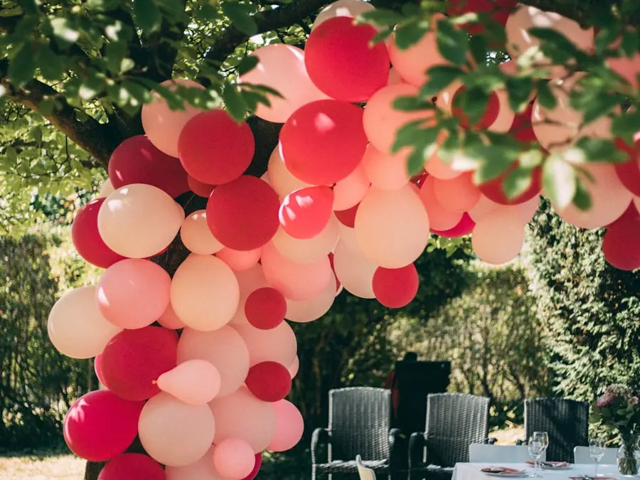 A pink and red balloon garland woven through a backyard tree over a party table